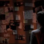 Man standing in front of investigator's corkboard in a dimly-lit room with his back to the camera.