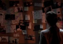 Man standing in front of investigator's corkboard in a dimly-lit room with his back to the camera.