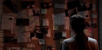 Man standing in front of investigator's corkboard in a dimly-lit room with his back to the camera.