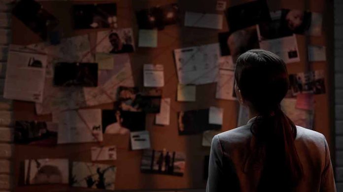 Man standing in front of investigator's corkboard in a dimly-lit room with his back to the camera.