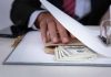 Hand concealing cash under a folder on a desk.