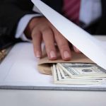 Hand concealing cash under a folder on a desk.