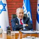 Man sitting at desk with Israeli flags behind.