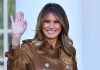 Woman smiling and waving in brown jacket.