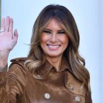 Woman smiling and waving in brown jacket.