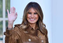 Woman smiling and waving in brown jacket.