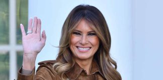 Woman smiling and waving in brown jacket.