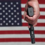 Hand holding gun against American flag backdrop.