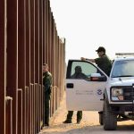 Border patrol agents near a tall metal fence.