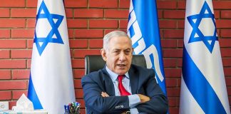 Man seated at a desk, Israeli flags behind.