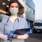 Doctor wearing mask and gloves, holding clipboard outside hospital.