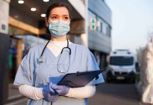 Doctor wearing mask and gloves, holding clipboard outside hospital.