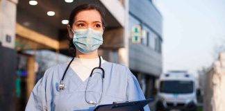 Doctor wearing mask and gloves, holding clipboard outside hospital.