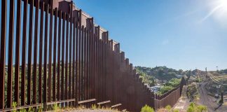 Tall metal border wall with rural landscape.