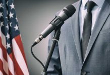 Man in a suit speaking into a microphone next to an American flag.