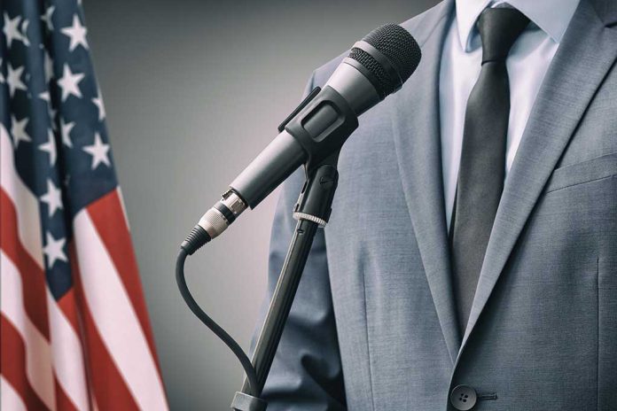 Man in a suit speaking into a microphone next to an American flag.