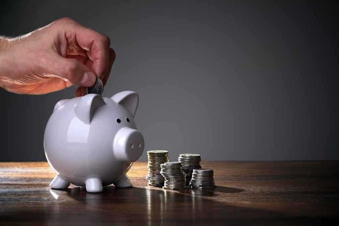 shutterstock_124029445.jpg A hand inserting a coin into a white piggy bank on a wooden table with stacks of coins nearby