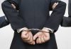 Person handcuffed in suit, escorted by two others.