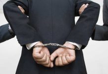 Person handcuffed in suit, escorted by two others.