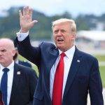 Man in suit waving while another man stands nearby.
