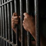 Prison MASSACRE Kills 35 – Suffocation Deaths Exposed Hands gripping prison cell bars tightly.