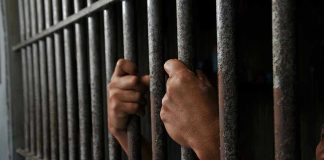 Prison MASSACRE Kills 35 – Suffocation Deaths Exposed Hands gripping prison cell bars tightly.