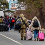 Group of people carrying luggage walking along a road with a soldier