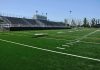 A football field with artificial turf and empty bleachers under a clear blue sky
