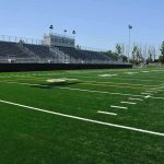 A football field with artificial turf and empty bleachers under a clear blue sky