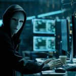Person wearing mask and hoodie at computer desk.