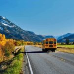 Teacher’s Disgusting Misconduct Rocks School! A yellow school bus driving on a rural road surrounded by autumn trees and mountains