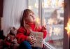 A child in a red sweater holding a gift while looking out a window decorated with lights