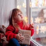 A child in a red sweater holding a gift while looking out a window decorated with lights