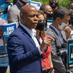 Adams Sides With Trump–Stuns Democrats Man speaking at campaign event with supporters behind him.