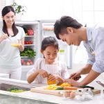 A father and daughter happily preparing vegetables in a bright kitchen, while a woman observes.