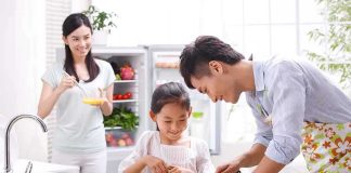 A father and daughter happily preparing vegetables in a bright kitchen, while a woman observes.