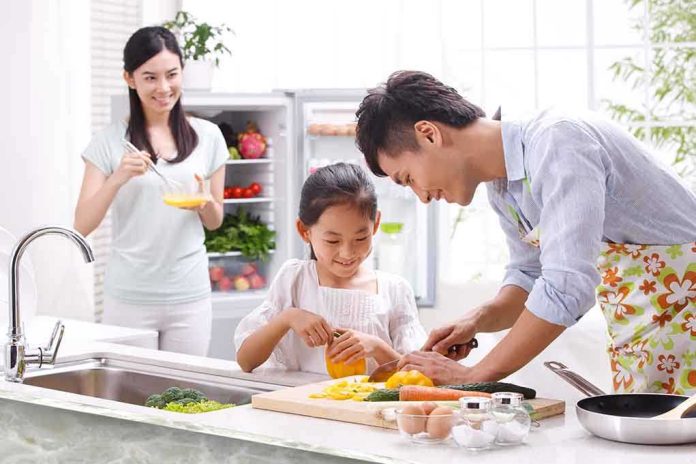 A father and daughter happily preparing vegetables in a bright kitchen, while a woman observes.