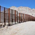 A long border wall stretches across a desert landscape with mountains in the background
