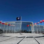 NATO Shakeup Looms — Europe’s Free Ride Ends Flags outside NATO headquarters building under clear blue sky.