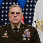 senior military officer in uniform with medals standing before U.S. flags