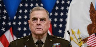 senior military officer in uniform with medals standing before U.S. flags