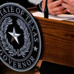 Close-up of the Texas governors seal on a wooden podium with hands gesturing in the background