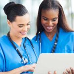 Fake Nurses Everywhere? Two nurses in blue scrubs smiling while looking at a tablet