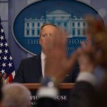 Press secretary at podium with raised hands in briefing room