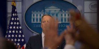 Chilling Iran Broadcast Names Barron Trump Press secretary at podium with raised hands in briefing room