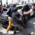 Masked Arsonists Ignite Themselves in Dispensary Attack Protesters surround and damage a police vehicle on a city street