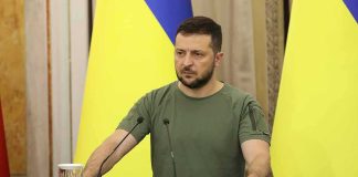 Zelensky Awaits Trump Envoys After Kremlin Huddle Man at a podium with Ukrainian flags