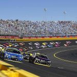 Stock cars racing on a banked track with grandstands in background