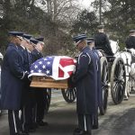 Military honor guard carries a flag-draped casket beside a hearse and caisson