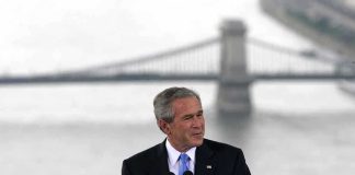 Bush Reopens 9/11 Playbook—What Got Unleashed? A man in a suit giving a speech at a podium with a bridge in the background